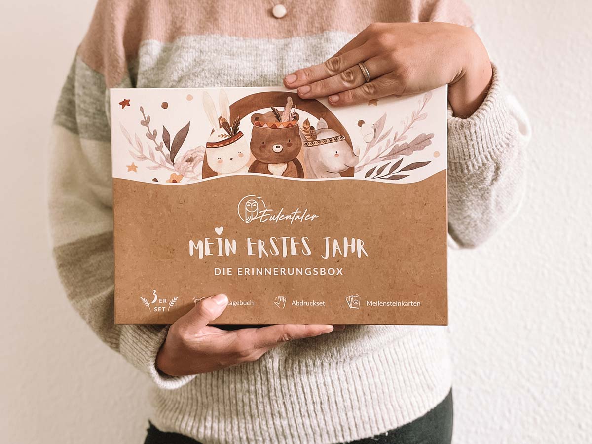 Nahaufnahme der Erinnerungsbox "Mein erstes Jahr" in den Händen einer Frau, enthält Babybuch, Abdruckset und Karten.